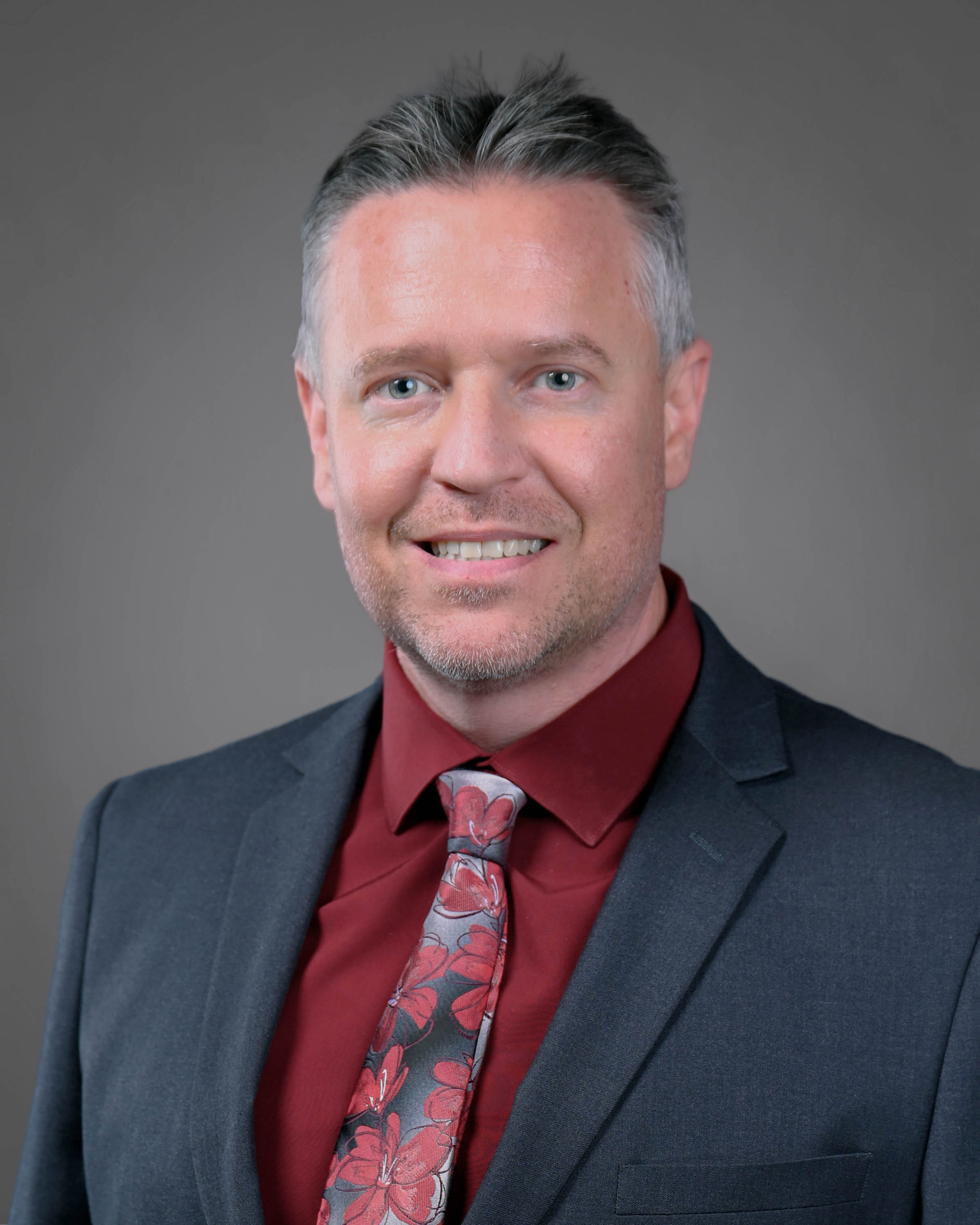 Smiling man with brown hair and short beard in a dark blue suit with a red shirt and tie.